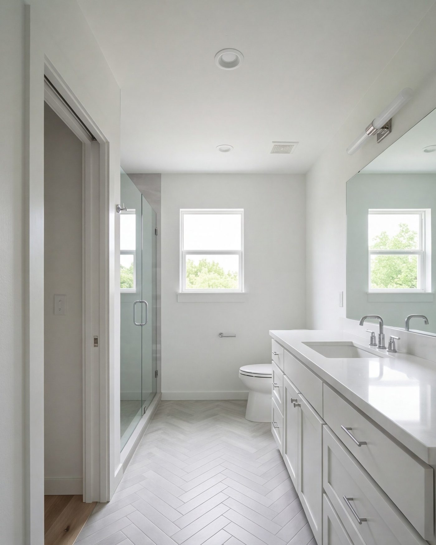 tall view into a finished hall bath from the hallway, glass door, wide vanity, light tile